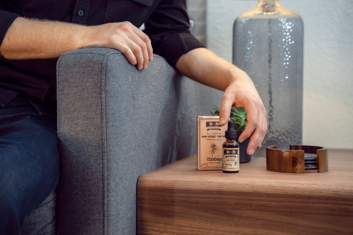 A man's hand reaches for a hemp oil tincture bottle on a wooden table indoors.